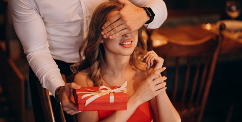 mężczyzna dłonią zasłania uśmiechniętej kobiecie oczy i wręcza czerwony prezent