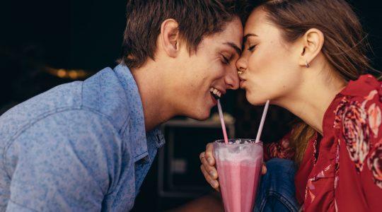 uśmiechnięta para pijąca razem shake truskawkowego, kobieta całuje mężczyznę w nos